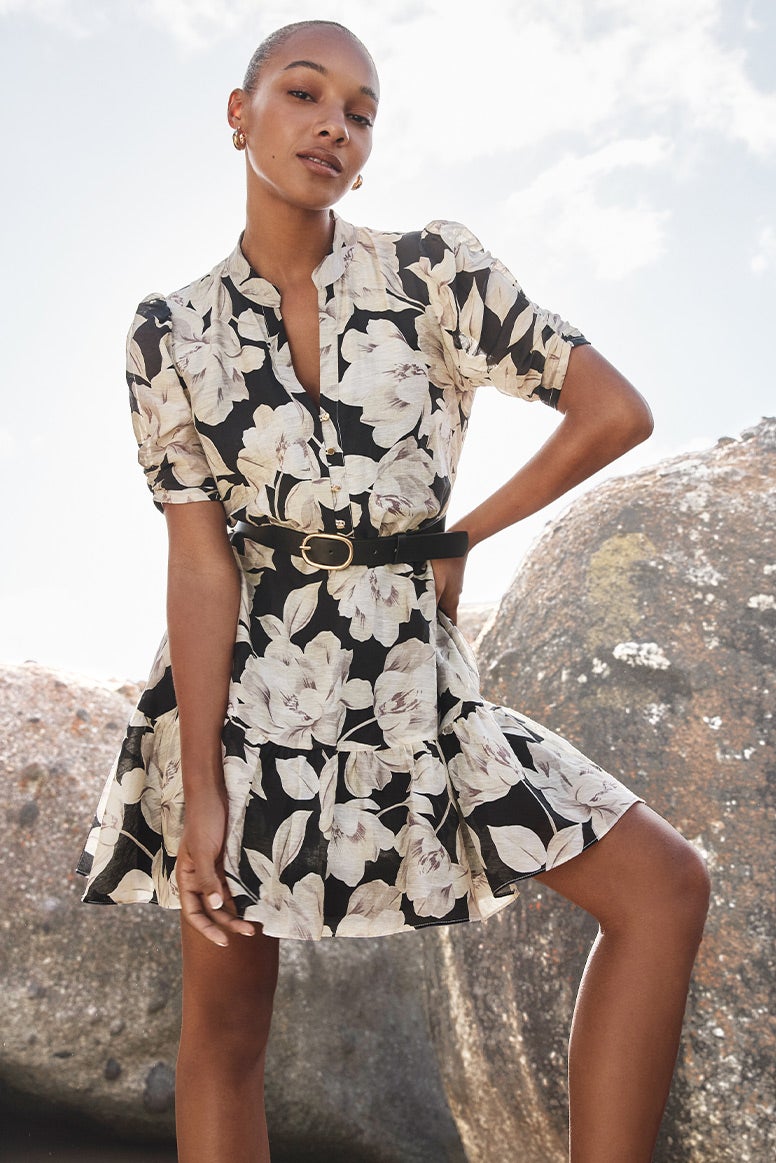 black model with slick back bun wearing a black and white floral mini dress with short sleeves and a black belt, she is posing in front of large grey rocks 