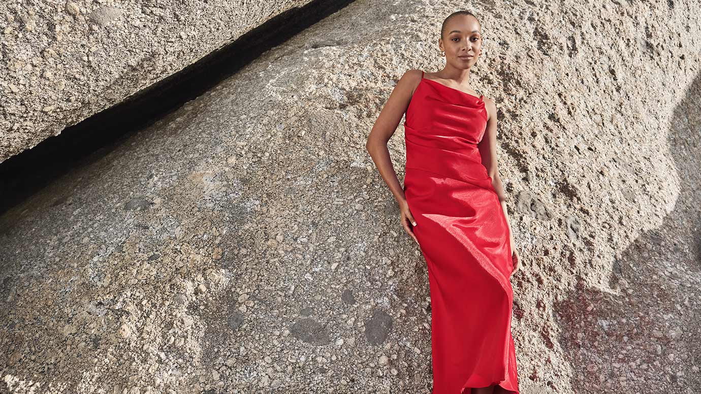 model with slick back bun wearing a strappy red slip style dress with gathered across the bust posing in front of large grey boulders