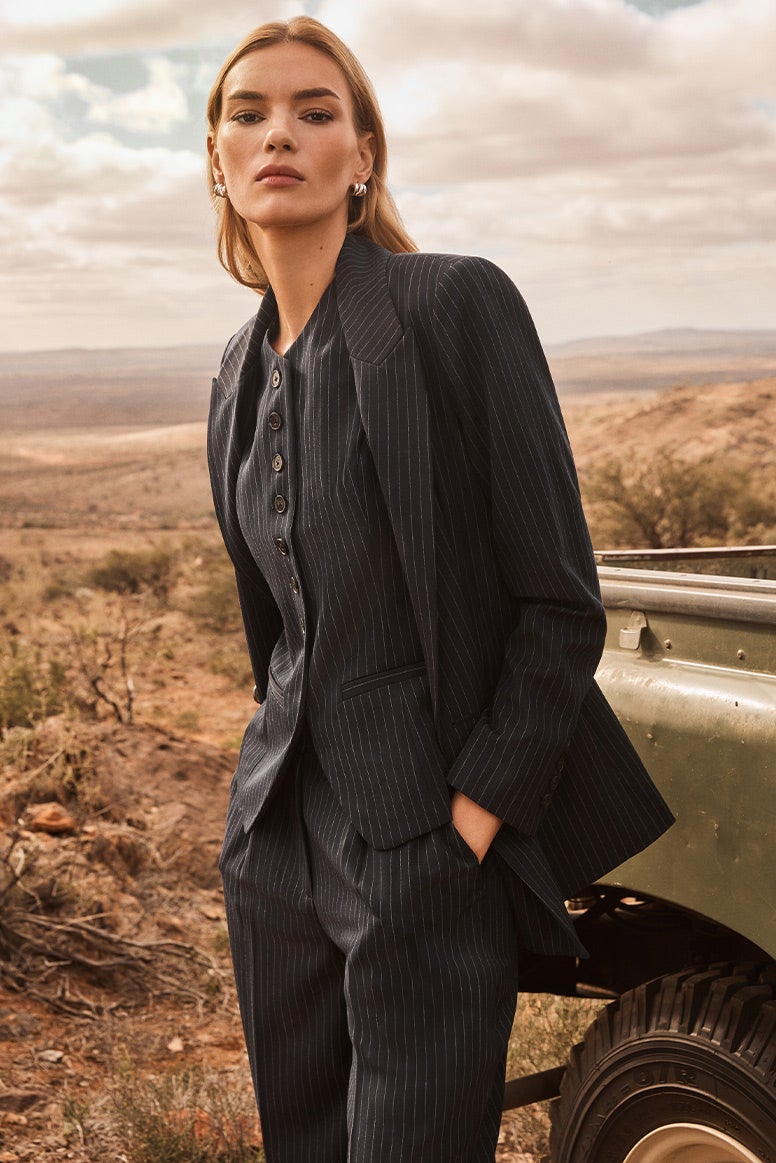 model wearing a three piece navy pinstripe suit standing near the back of an old green vintage pickup truck, she has her hands in her pockets, the landscape behind her is dusty and remote