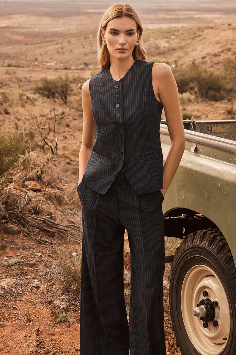 model wearing navy pinstripe pants and a matching, button up, longline waistcoat is standing near what looks to be a safari truck in a dusty remote landscape, her hands are in her pockets and she is staring directly into the camera