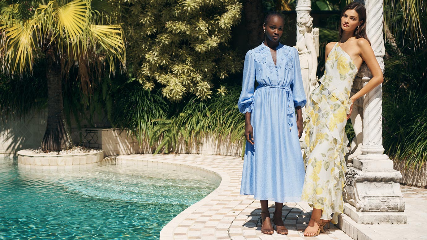 two models wear summery outfits gathered at the edge of a simple and chic pool with a manicured gardens and grand architecture.