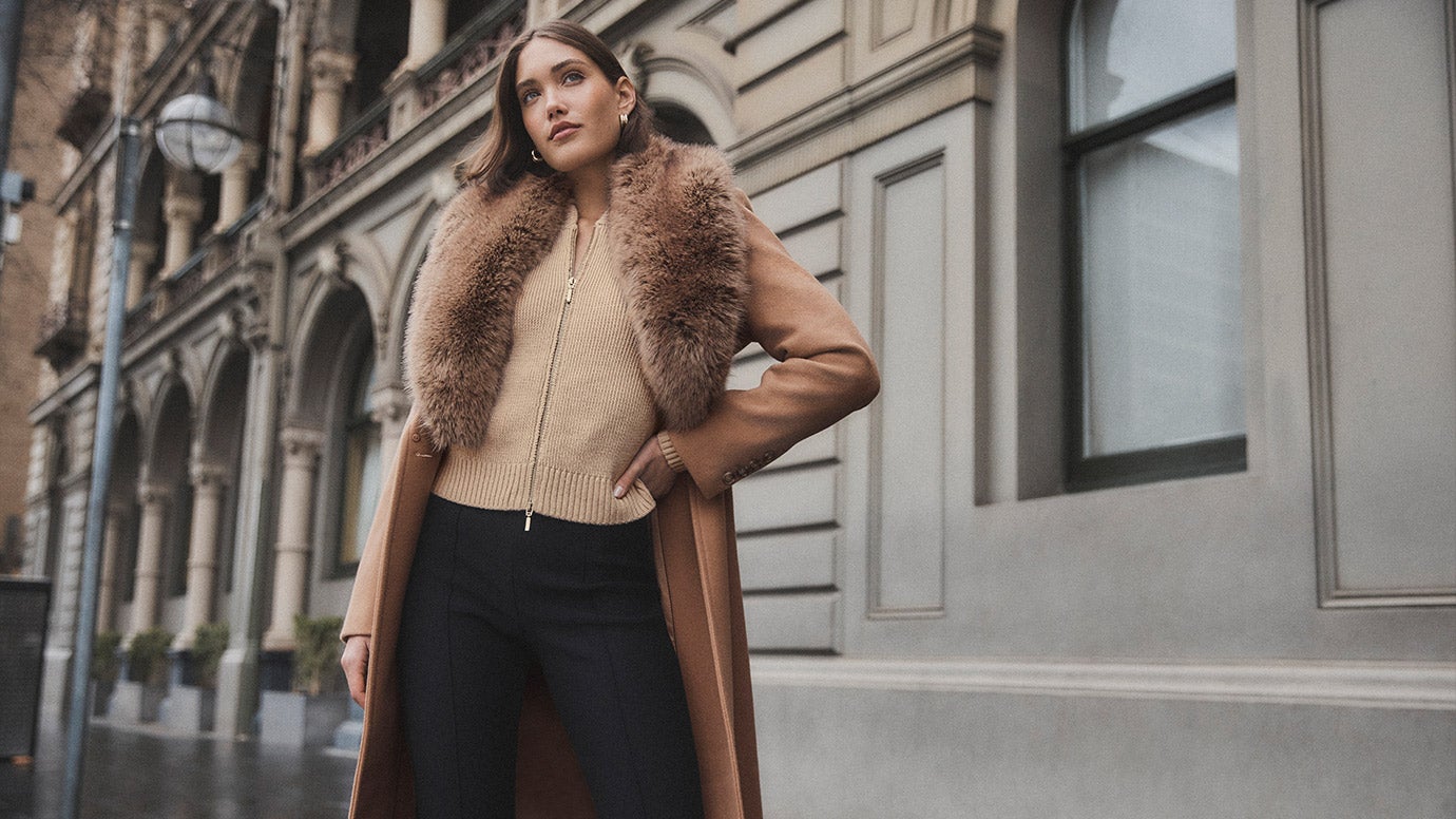 model with a short brown bob wearing camel coat with big faux fur collar trim, jeans, and a light camel jumper with a silver zip down the front poses in an empty side street with a grey building in the background, one hand is on her hip
