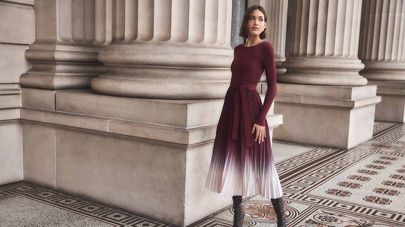model with brown bob wearing a dress that looks like a long sleeve knit top in plum with a pleated plum and white ombe skirt and tall brown boots, she poses near giant old pillars and a patterned paved floor