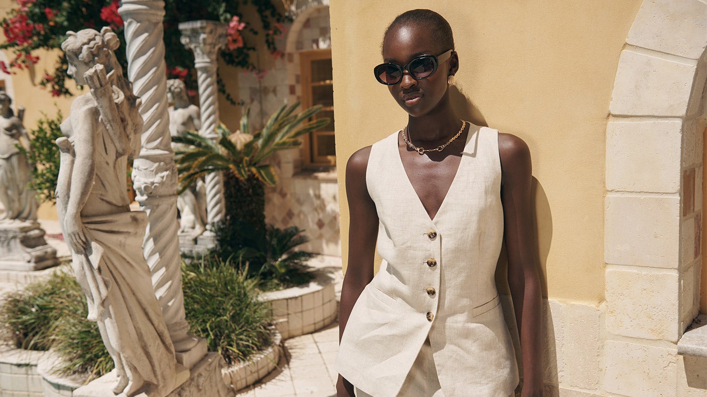 Model wears dark sunglasses and neutral tone waistcoat with matching suit pants. She leans against a sun-drenched yellow wall surrounded by red flowers and the architecture of Palazzo Rosa.