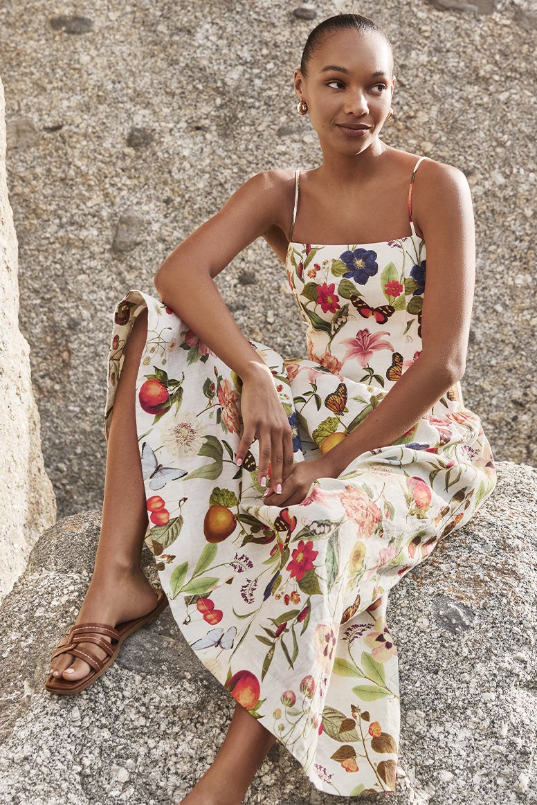 black model with slick back bun wearing a strappy floral sundress, the print is colourful and includes flowers, butterflies, and oranges
