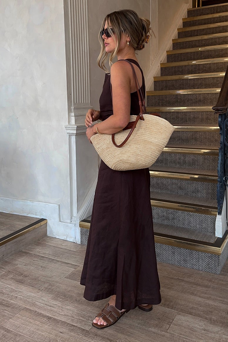 influencer tanis rose wearing a long flowing linen dress in deep chocolate brown, she is standing in profile turned to the left in front of a staircase, she is wearing slides, dark sunglasses, and is holding a straw tote 