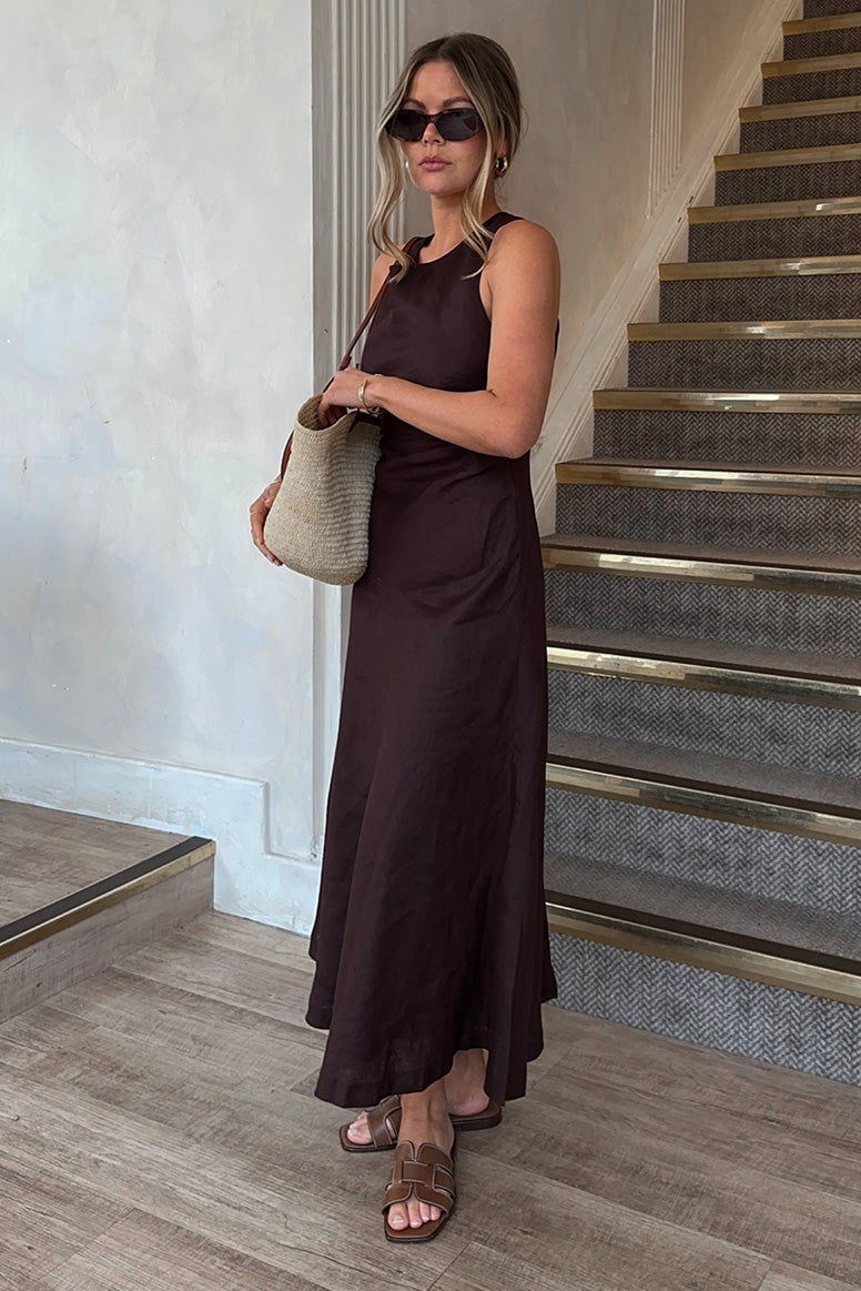 influencer tanis rose wearing a long flowing linen dress in deep chocolate brown, she is standing in front of a staircase, she is wearing slides and dark sunglasses and is holding a straw tote 