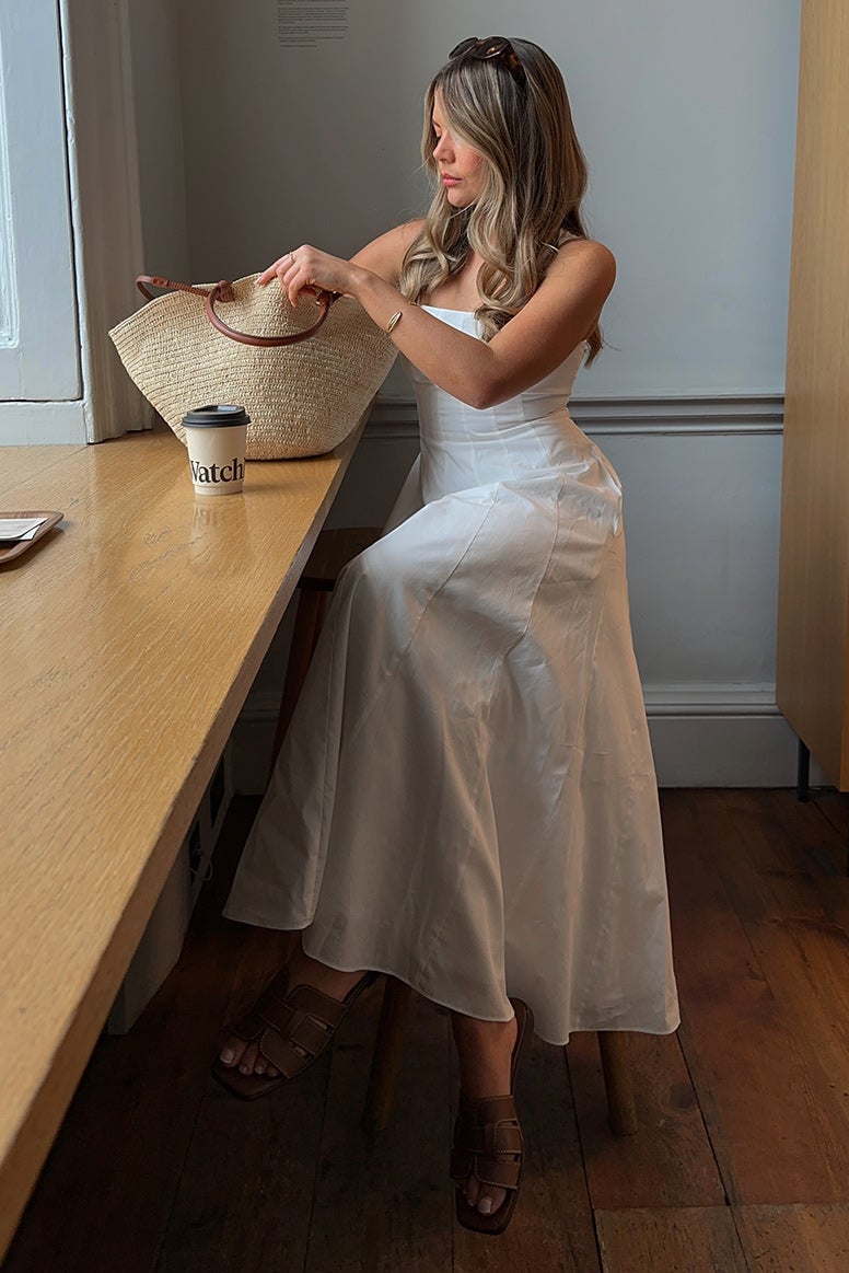 influencer tanis rose wearing a long flowing white dress with a seamed bodice, she is sitting at a simple wooden bench facing an open window in a cafe, she is looking into her straw tote which is placed on the counter in front of her
