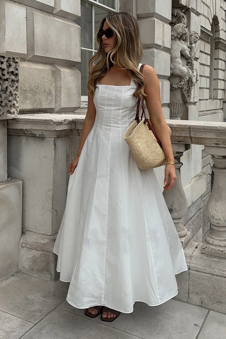influencer tanis rose wearing a flowing white dress with seamed bodice and wide straps, she is standing near an old stone building and small pillars are seen behind her, she is wearing dark sunglasses and is holding a straw bag