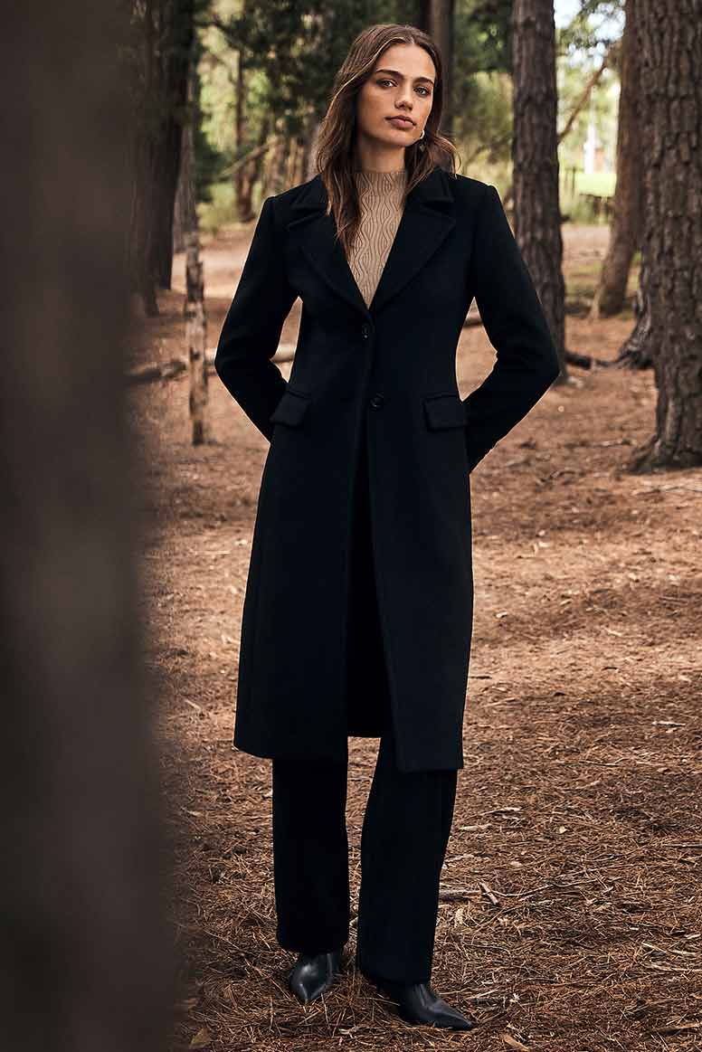 a model wearing a long black coat, a camel coloured top, and black pants with pointed boots poses in a forest setting, a tree trunk is in the foreground to the left