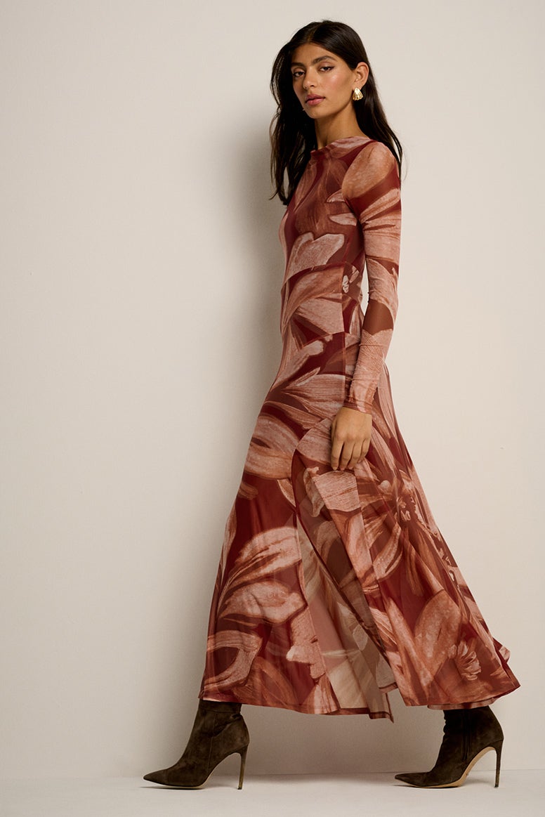 a model with long dark hair wears a rust floral dress with long sleeves and a long softly pleated skirt with choc brown tall boots, she is poses as if she is caught midwalk