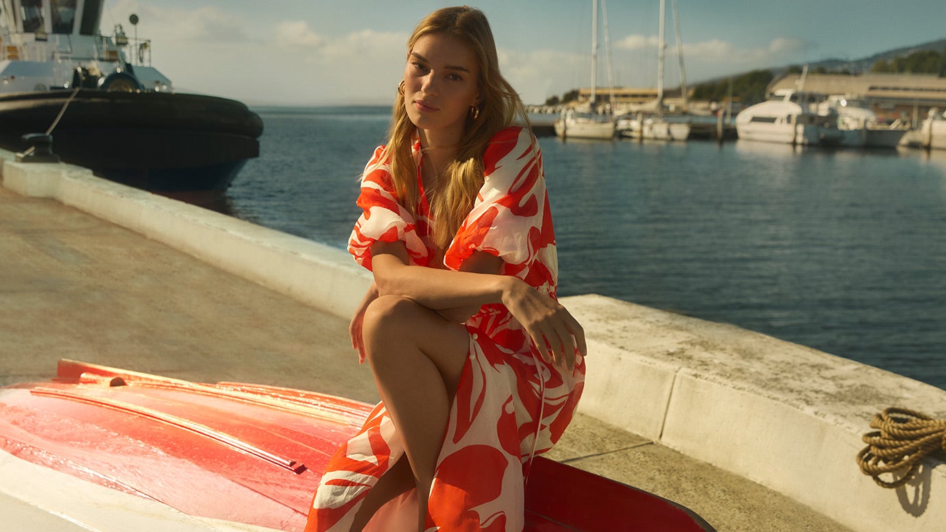 model sits on an upturned red and white rowing boat, she is wearing a long blue denim dress with buttons down the front and wide tank straps, the water and large boats are in the background