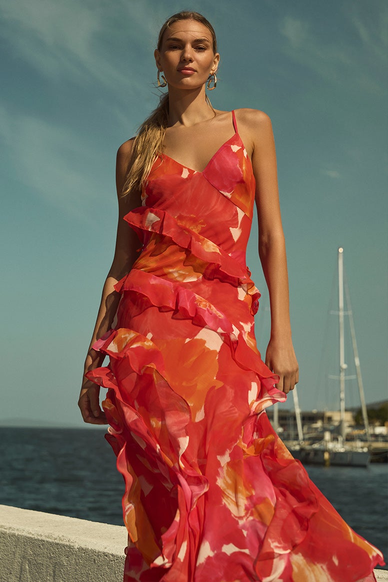 model wearing a strappy dress with ruffle trims in a vibrant tropical floral print in shades of red pink peach and white, you can see a blue bay behind her with large boats docked at a wharf