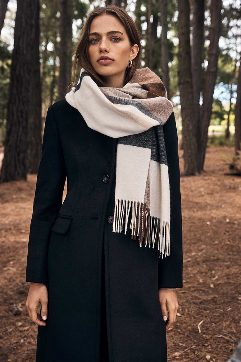 a model poses in a forest wearing a big check scarf in tones of cream and browns and a long black coat 