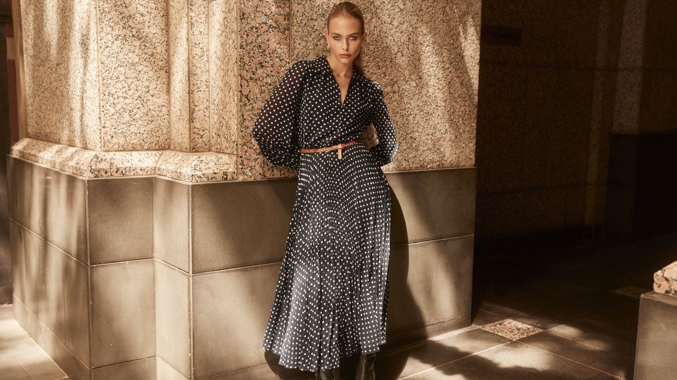 model leaning against a sandstone building in dappled sunlight wearing a long polka dot belted dress with long blouson sleeves