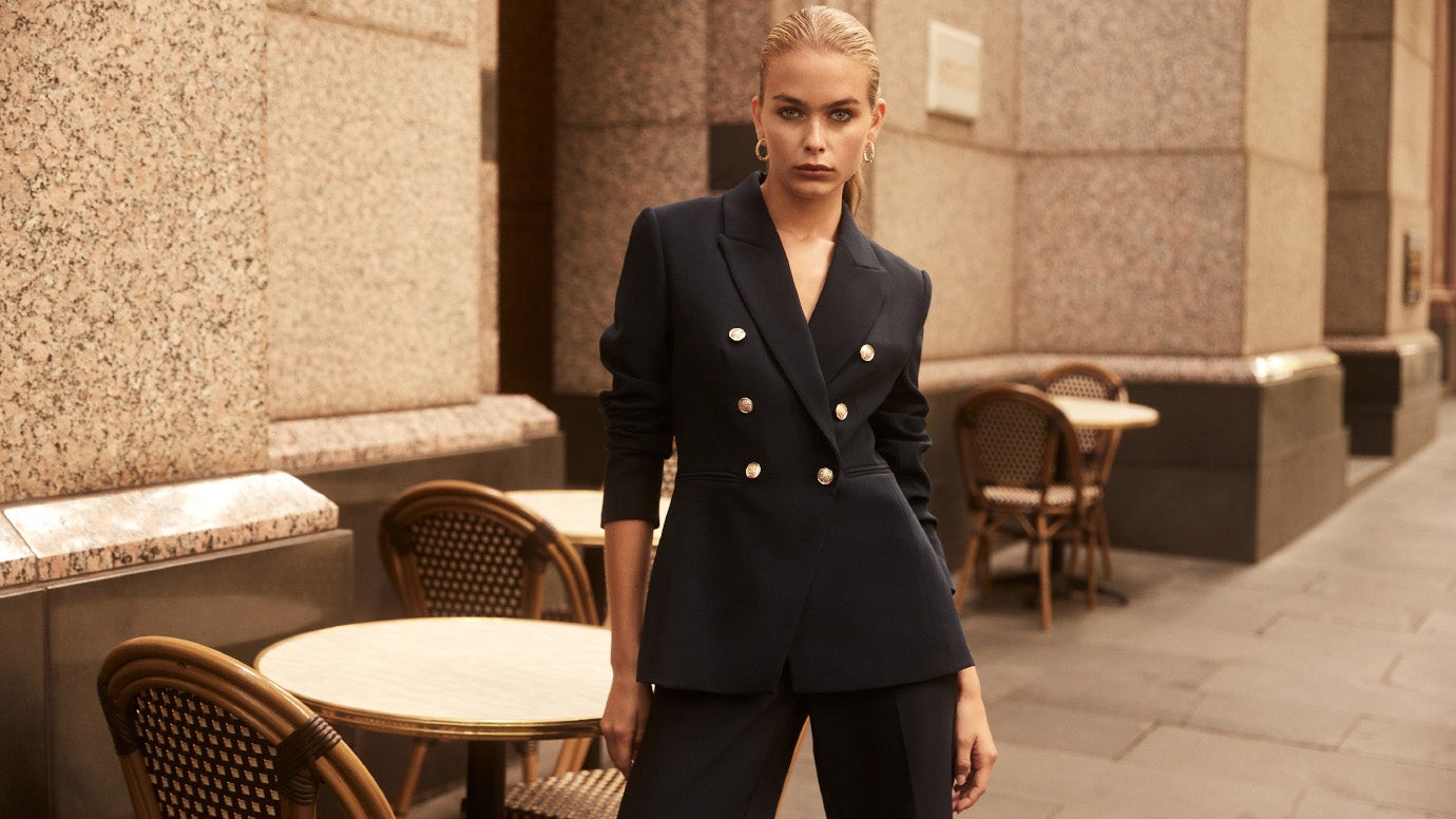 blonde model posing in front of french cafe style tables and chairs in a black suit with gold tone buttons