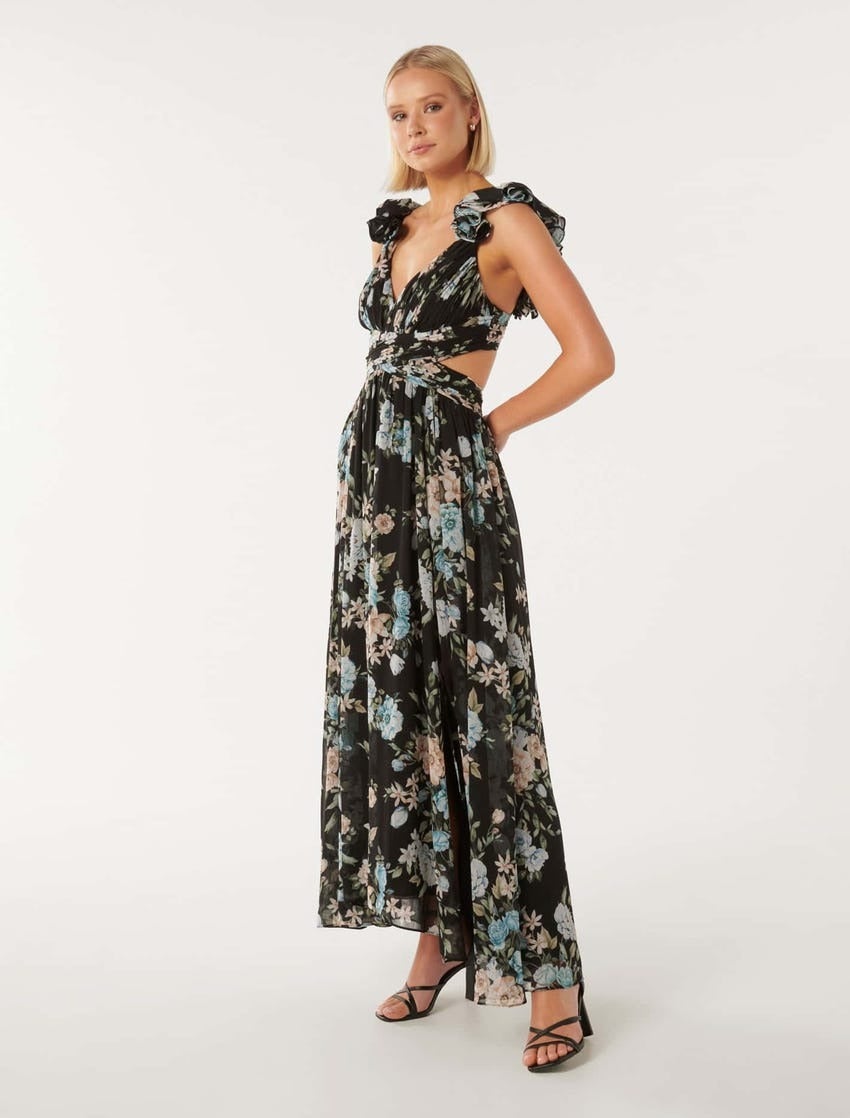 model with a blonde bob posing with a hand on hip wearing a long flowing black and blue floral dress with ruffle cap sleeves and cut outs at the side bodice