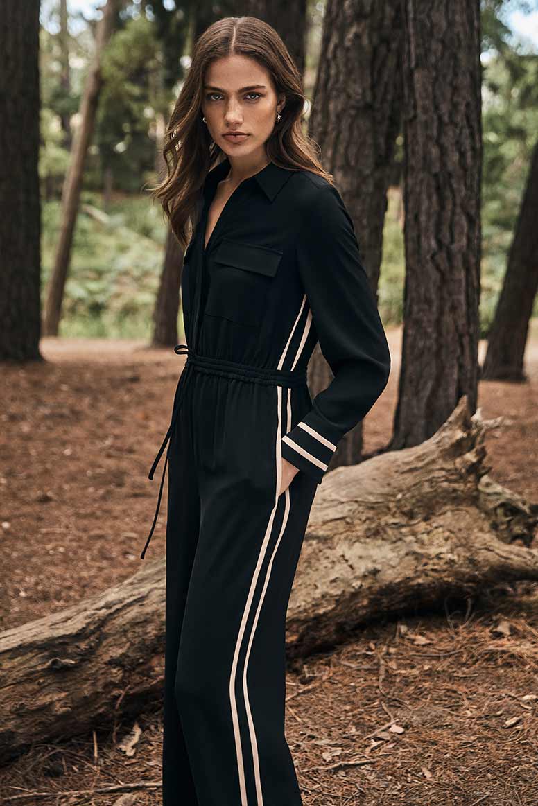 a model poses in in a forest wearing a black jumpsuit with a racing stripe down the sides in camel, she has her hand in her pocket