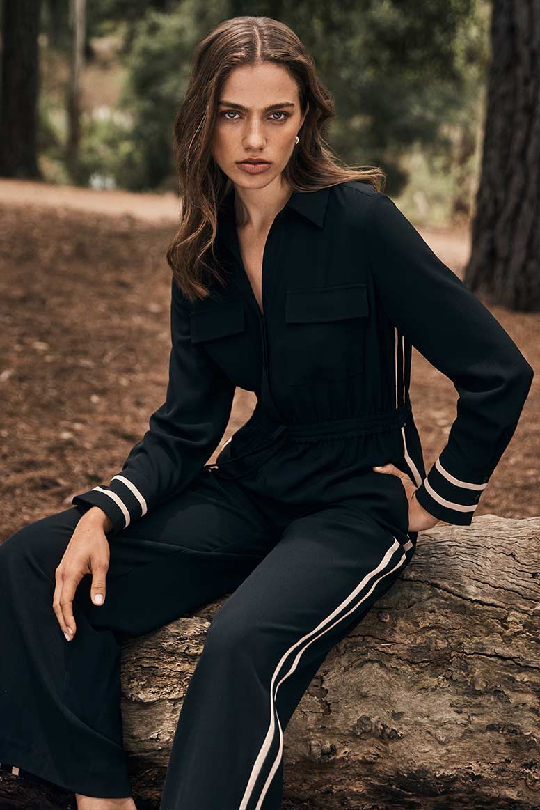 model poses in a forest setting, she sitting on a fallen log, she is wearing a long sleeve jumpsuit with a racing stripe detail, it is black with a camel coloured stripe