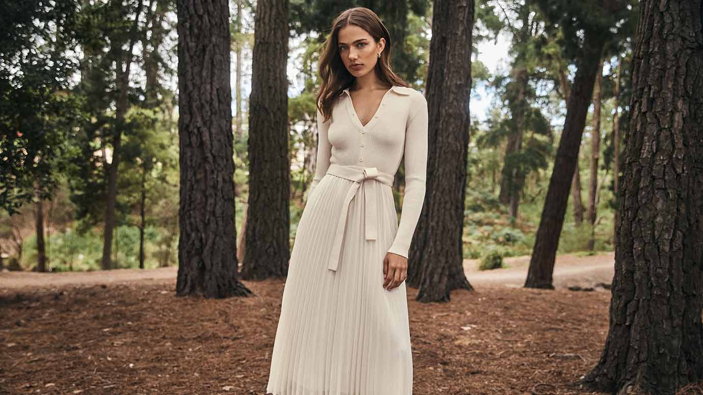 a brunette model is wearing a cream dress where the bodice looks like a knit polo sweater and the bottom of the dress is a pleated full skirt made from viscose, she is poses in a forest