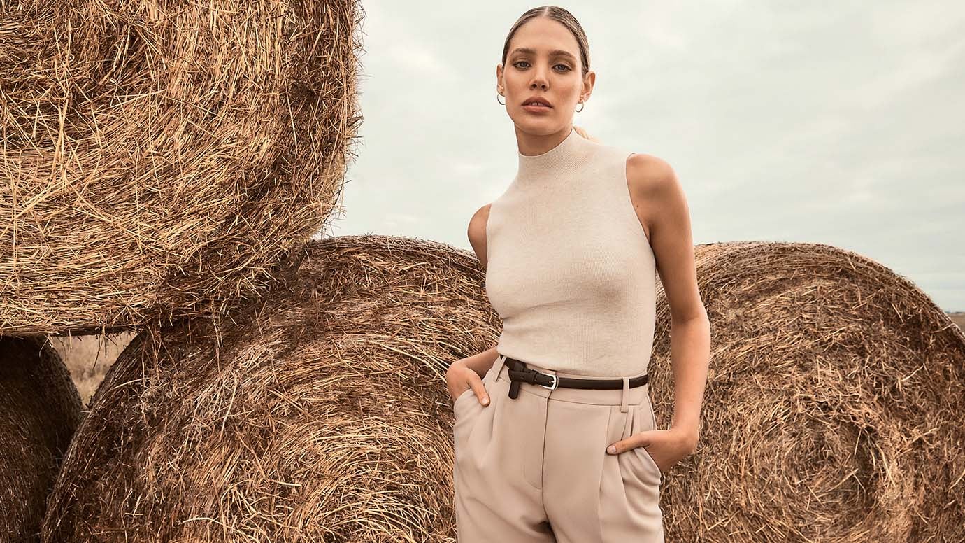 a model with slicked back hair posing in front of rolled hay bales wearing a stone coloured high-neck knit tank with stone coloured pants and a thin black belt 