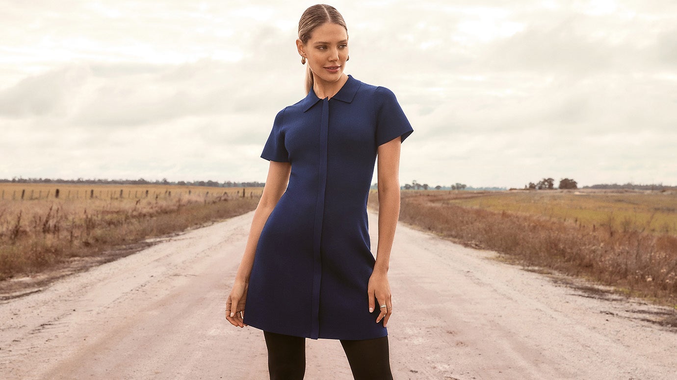 model with slicked back ponytail wearing a minimalist navy knit dress poses on a wide open road with empty fields on both sides