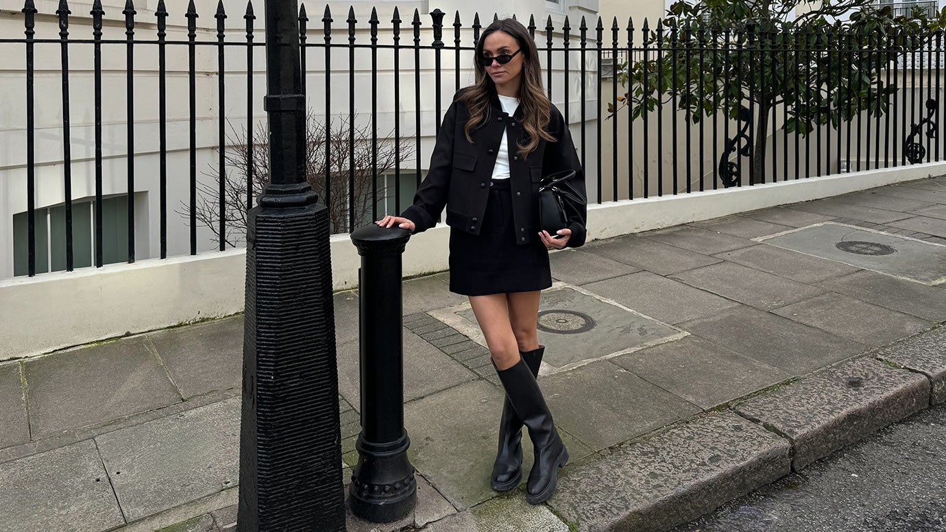 influencer felicity bird standing in front of a large cream building and black iron fence dressed in knee high black boots black mini skirt white tee and black bomber jacket she is wearing black sunnies