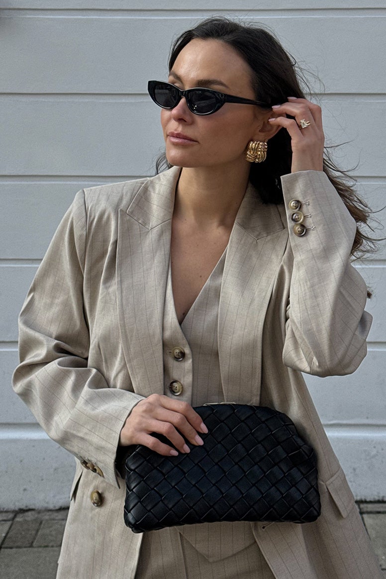 influencer felicity bird wearing a beige/grey pinstripe waistcoat and blazer holding a black woven pouch bag she is tucking her long hair behind her ear, you can see she is wearing a large gold earring and is wearing black sunnies