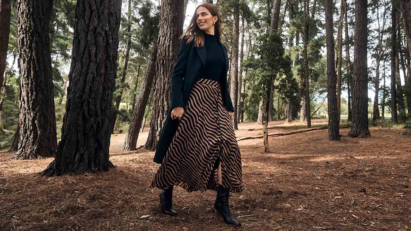 brunette model wearing a black top and coat with a long satin wrap skirt with a bold chocolate and black stripe print, she is standing in a forest looking to the left