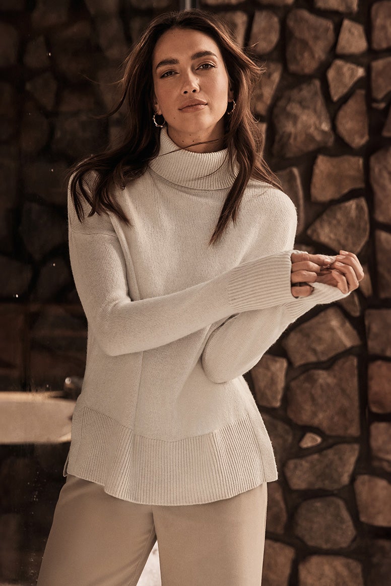 a brunette model poses in front of a rock wall or column wearing camel trousers and a cream roll neck jumper, you can see a few small snowflakes in the air behind her