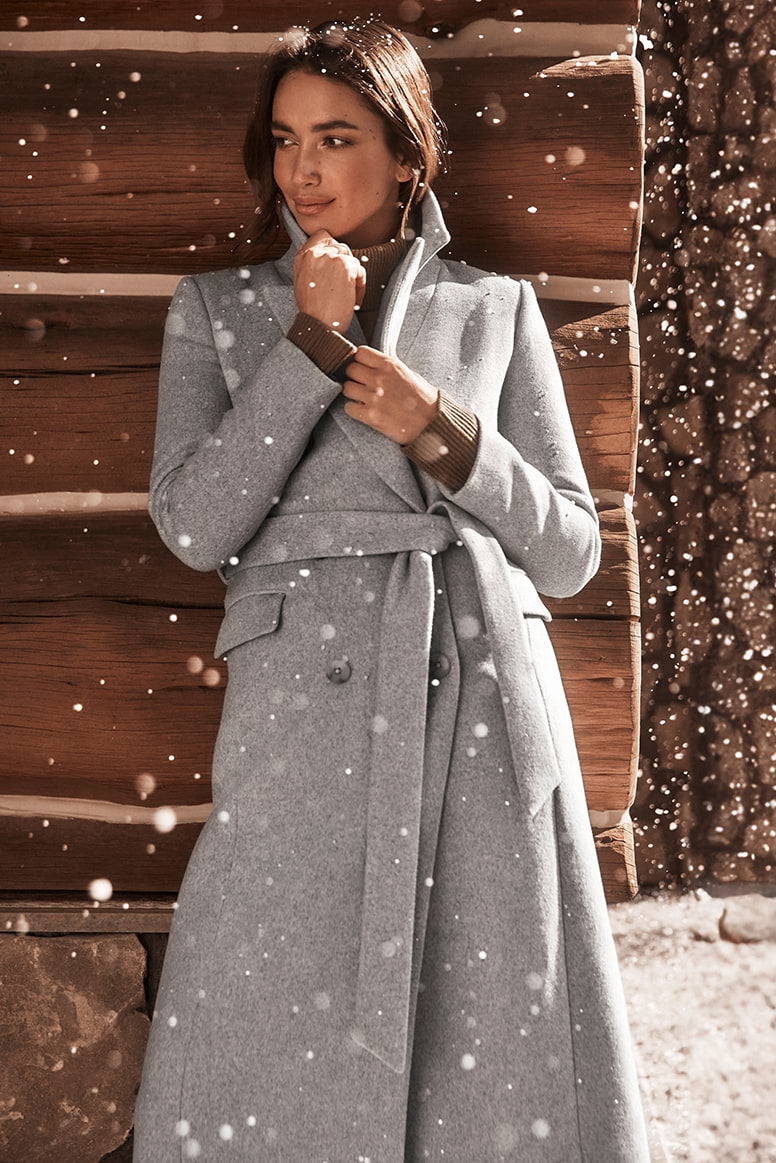 model with brunette hair wearing a long grey belted coat, holding it closed at the bust, you can see she is wearing a brown jumper underneath, she is leaning against a log cabin as snow falls around her