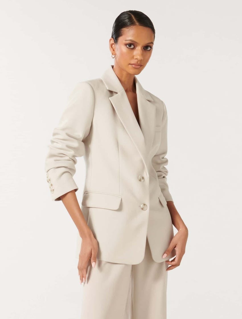 model with slick back brunette hair wearing a stone coloured suit with the arms rolled up with a plain grey background