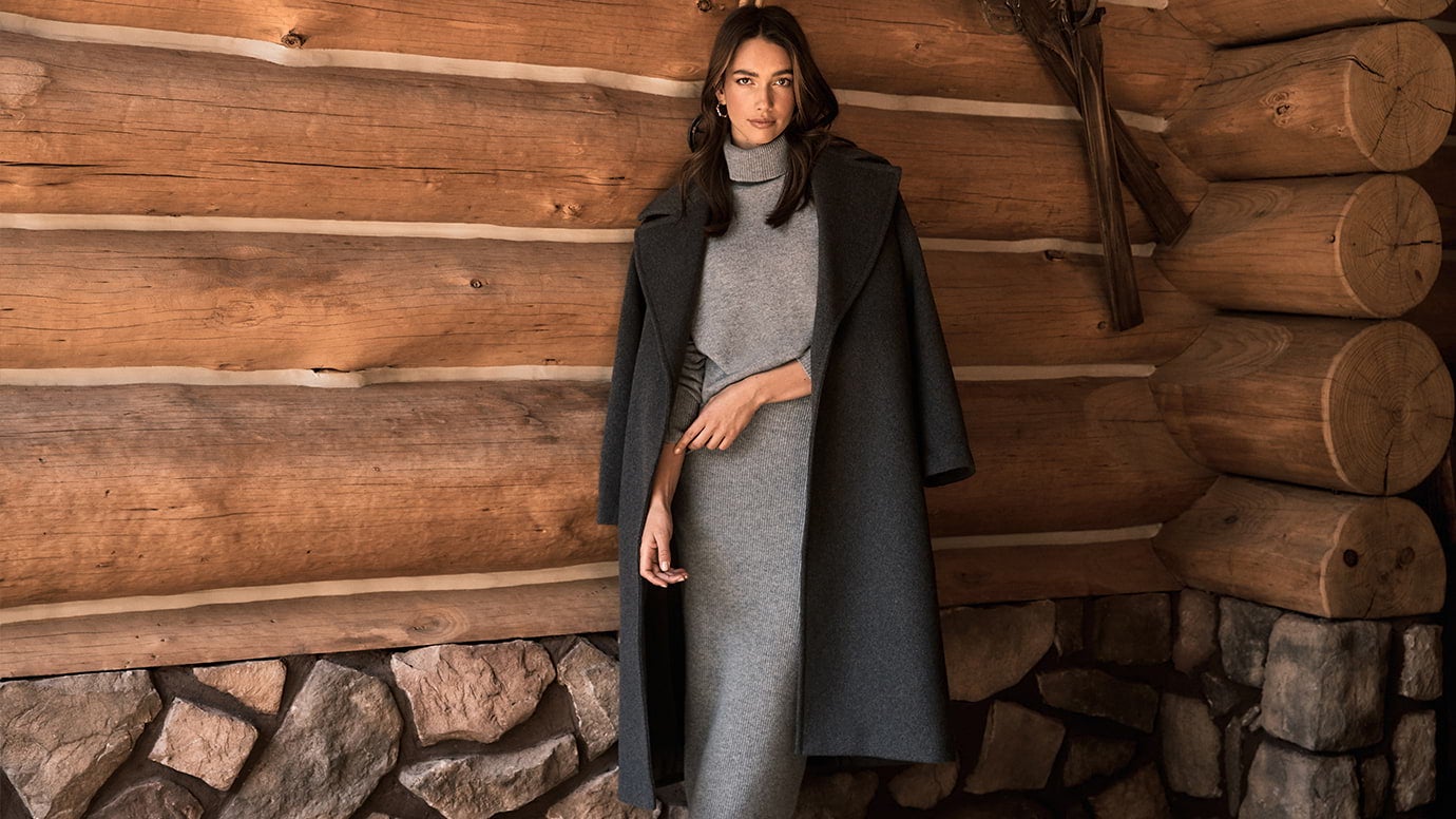 a brunette model posing in front of a log cabin wall wearing a long charcoal coat with a lighter grey knit set on underneath, she is crossing her arm against herself